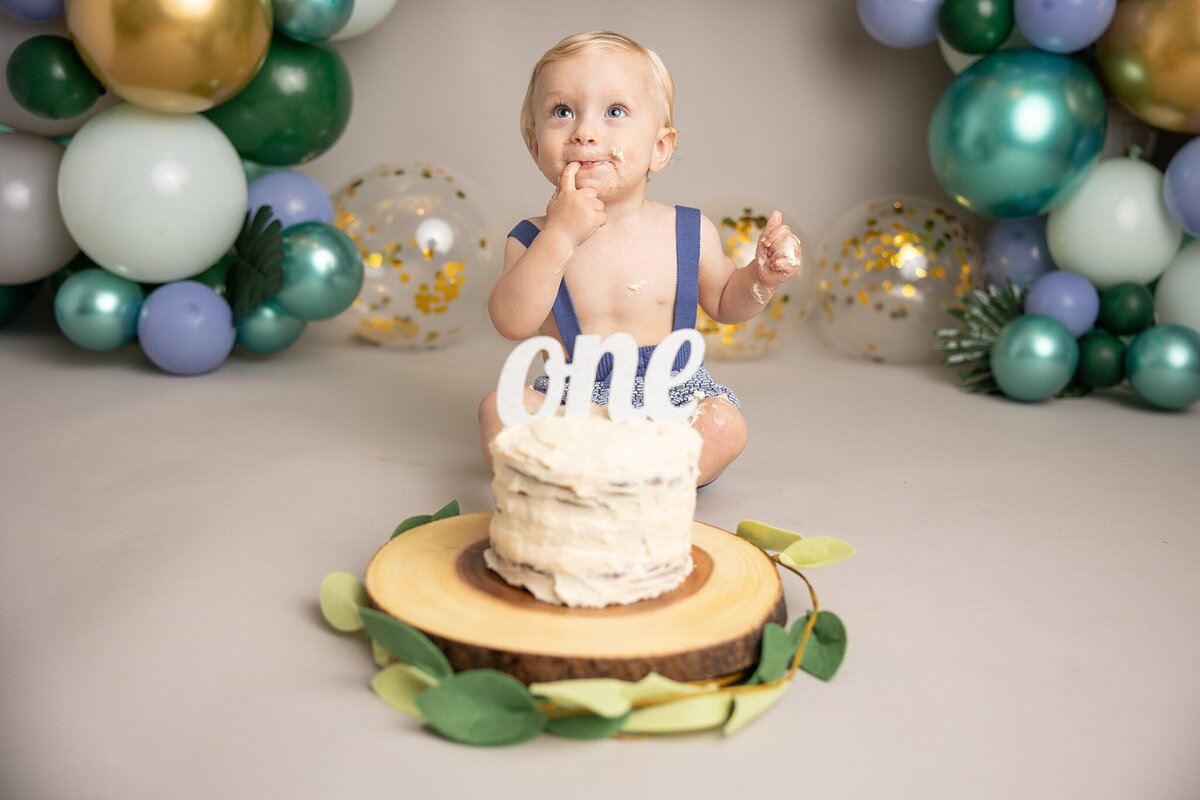 baby boy in front of balloon garland sitting behind one cake for first birthday pictures