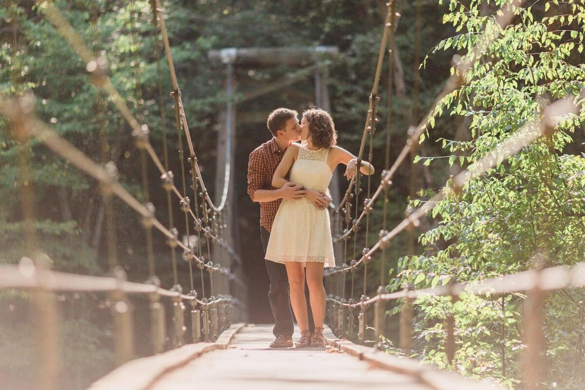 Romantic scene on a suspended bridge in the woods, with a couple kissing and sunlight streaming through the trees.