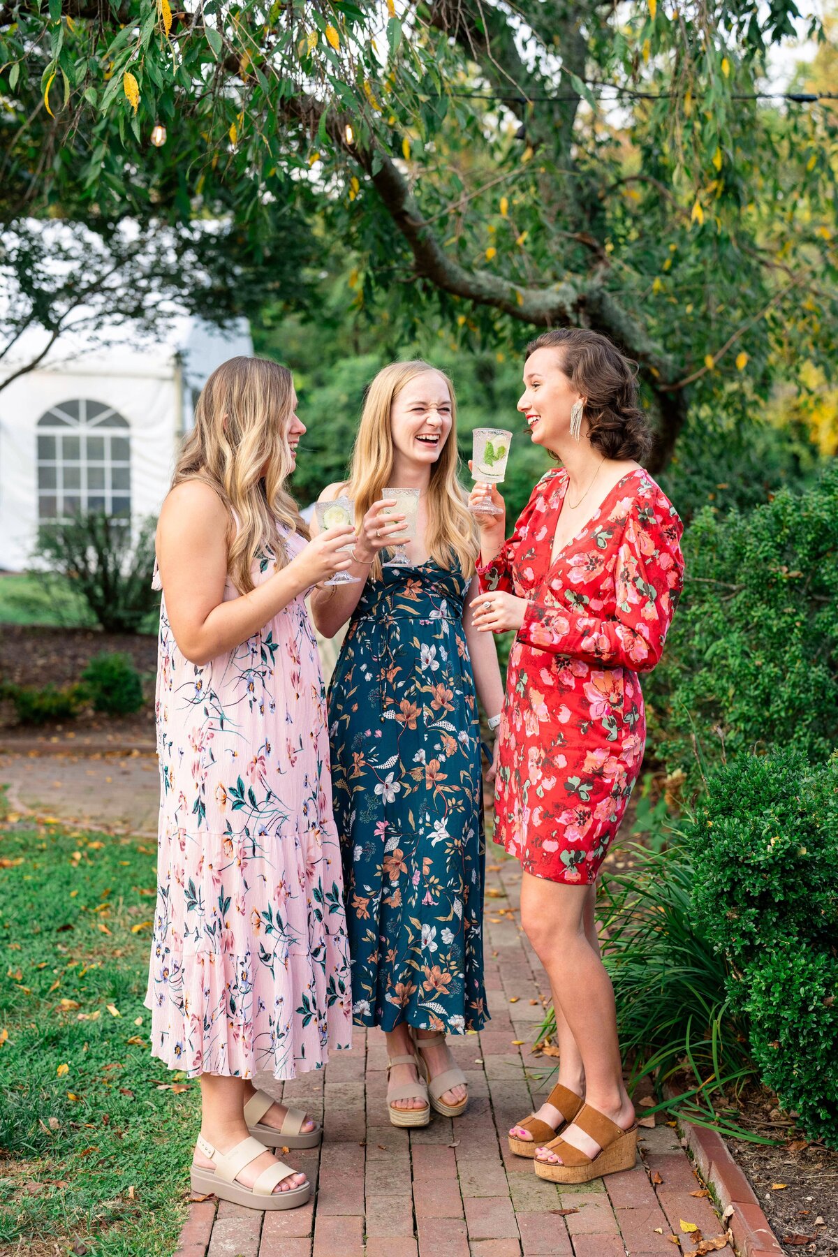 Three women in floral dresses stand on a brick path, laughing and holding drinks, surrounded by greenery. A tree with hanging branches and a white tent are visible in the background.