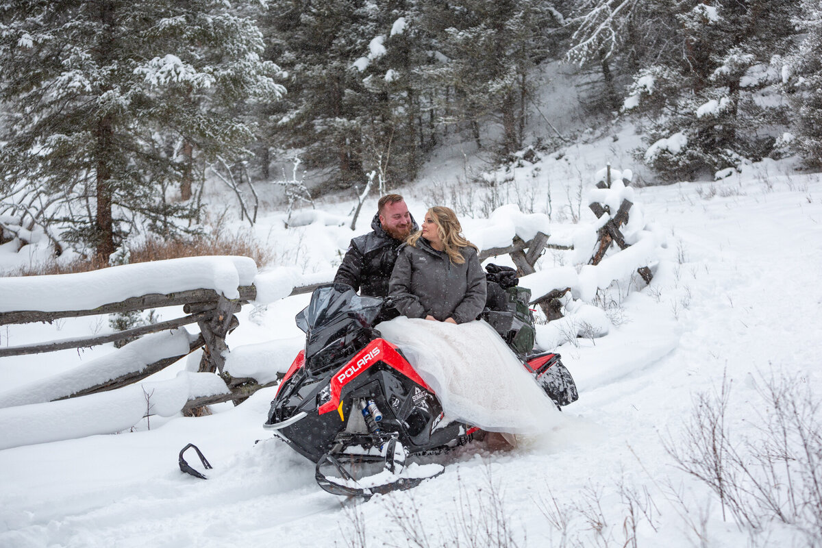 A bride sits on a snowmobile on her wedding day while her groom stands in the snow behind her.