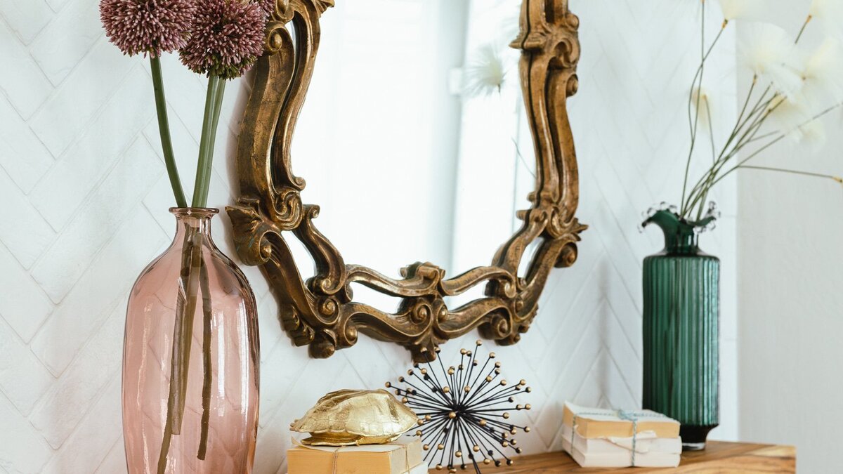 Elegant interior design vignette with vintage gold mirror, pink glass vase, and modern accessories styled on a wood mantel