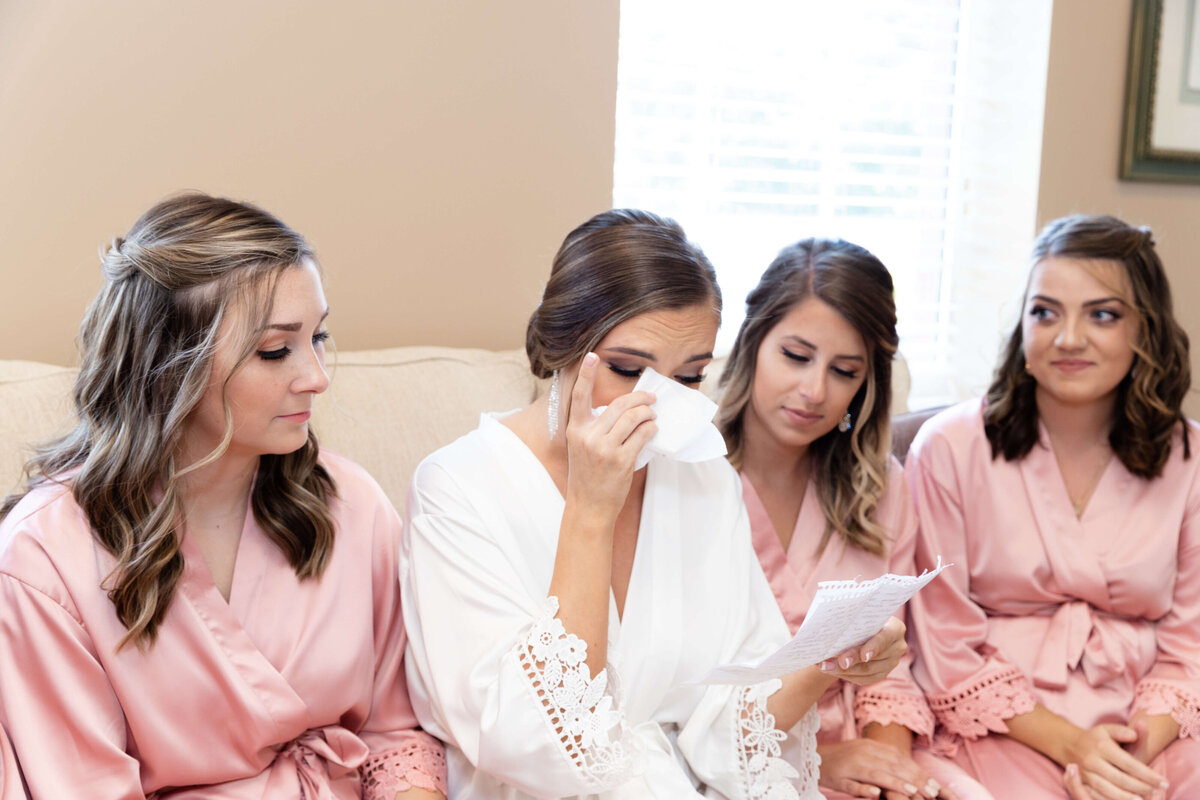 Bride wiping eye with tissue as she reads note with bridesmaids sat beside her