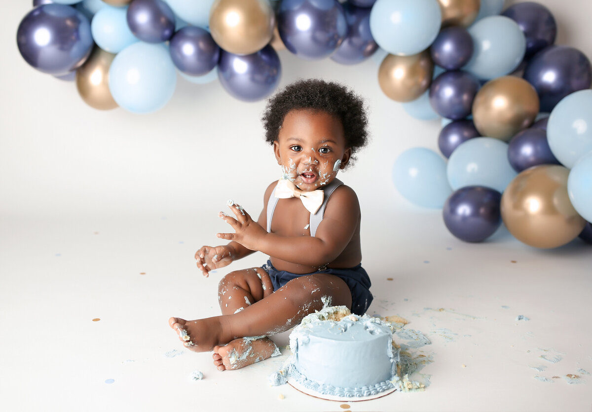 Birthday boy cake smash at top West Palm Beach, FL cake smash photography studio. Black baby boy wearing diaper cover, suspenders, and a pink bow tie is sitting behind a pale blue iced cake with icing on his legs and face.  In the background, there is a blue, gold, and navy balloon arch.