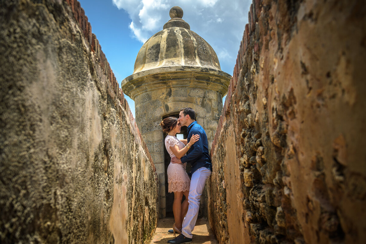 A couple shares a loving embrace near a historic stone structure under a blue sky.