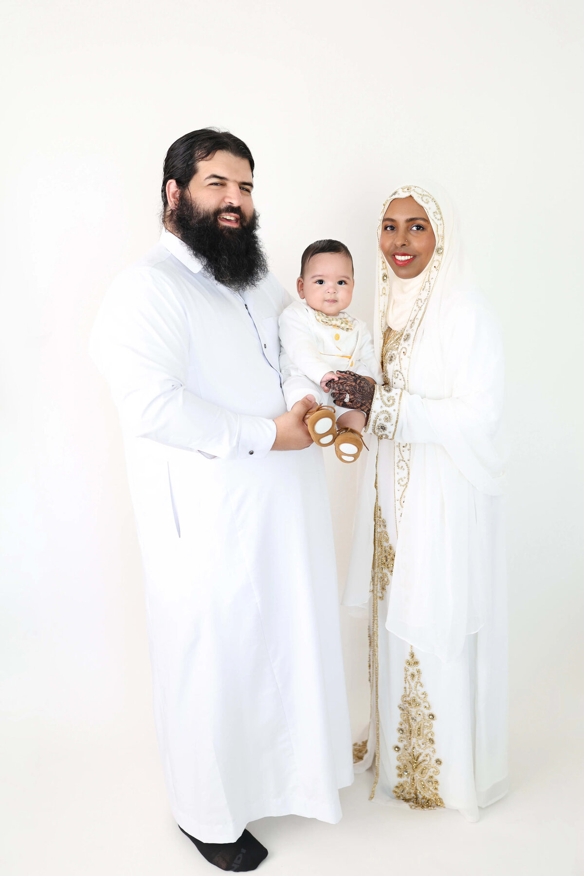 Family dressed in traditional white and gold attire holding baby in studio - cultural family session in Maple Grove