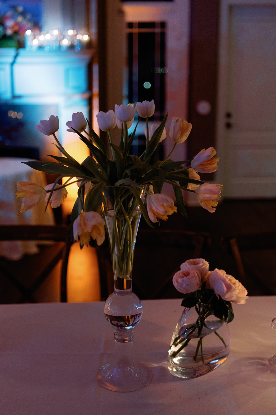 Elegant white tulips and soft pink roses in glass vases on a reception table, softly lit with warm amber and cool blue uplighting at Callie & Hayes’ Burritt on the Mountain wedding reception.