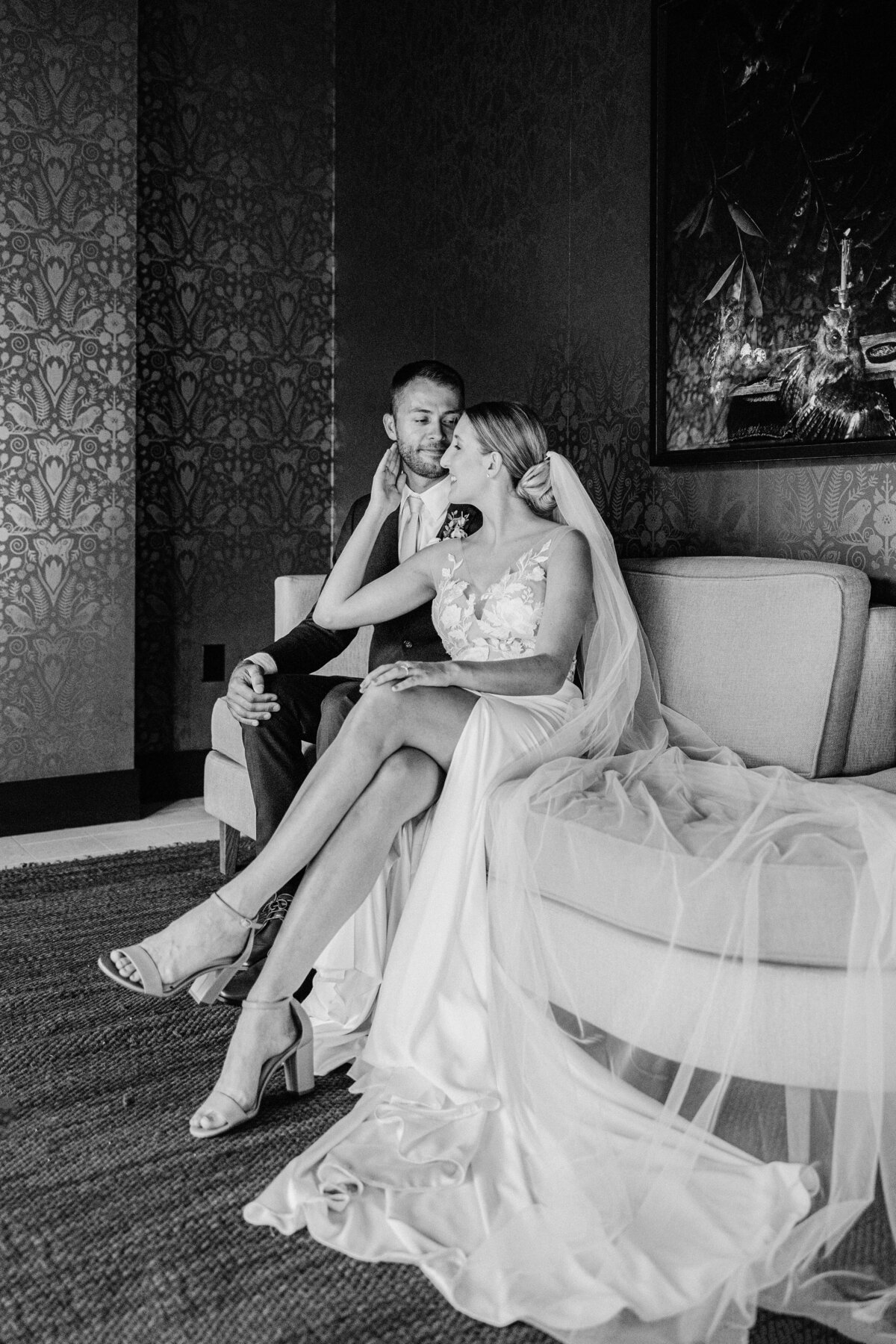 Bride in a modern floral-appliqué gown gently cups the groom’s face while seated on a curved sofa at The Alida Hotel, Savannah—captured by luxury wedding photographer Amia Marcell.