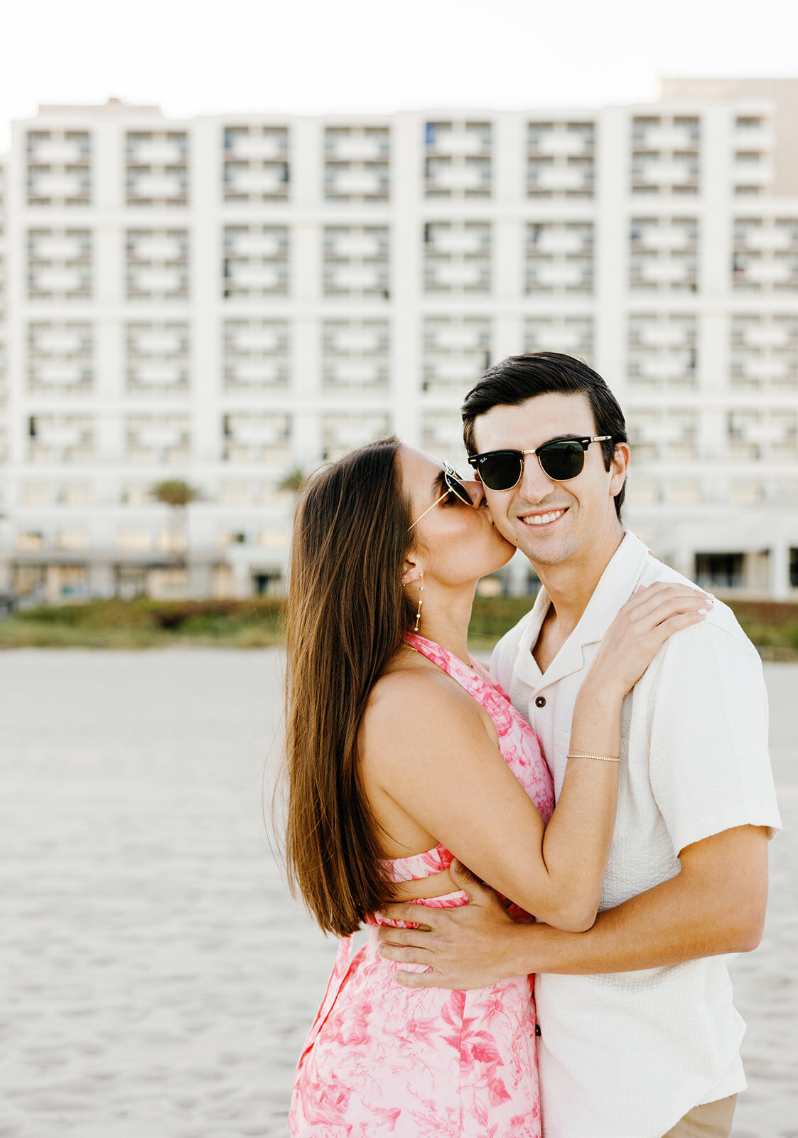 Rebecca, in a stunning floral dress, leans in to kiss Graysen in front of a modern high-rise at Marriott Hotel Fort Lauderdale, creating an elegant and stylish engagement portrait.