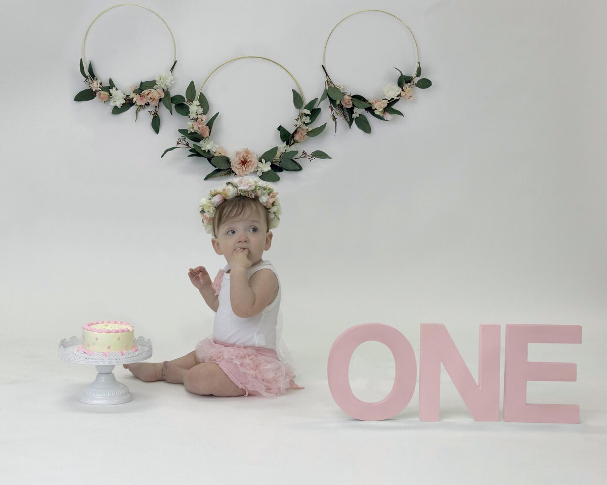 An Orange County milestone photography image of a baby sitting in a studio in a pink tutu eating a birthday cake with flowers behind her