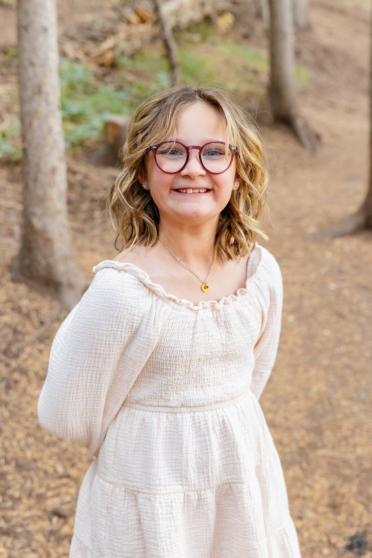 A young girl with glasses and curly hair beams at the camera, dressed in a cream-colored dress, standing confidently in a wooded setting at Tibble Fork.