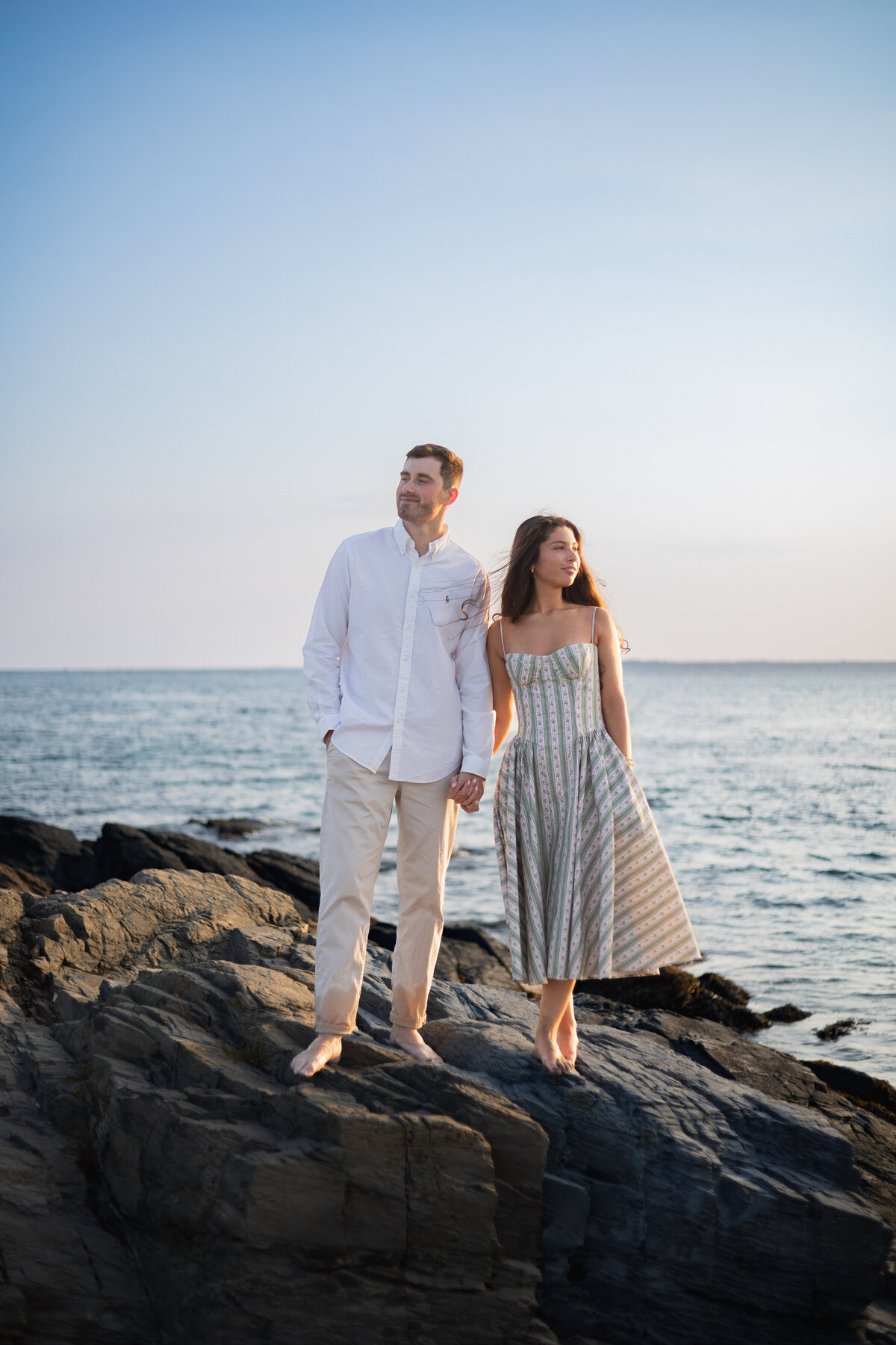 Collins Beach Newport RI | Kelsey Sheehan Photography Timeless Rhode Island Weddings | A couple, barefoot and holding hands, walks on seaside rocks against a serene sunset sky. The man wears a white shirt; the woman, a striped dress. Romantic and peaceful.