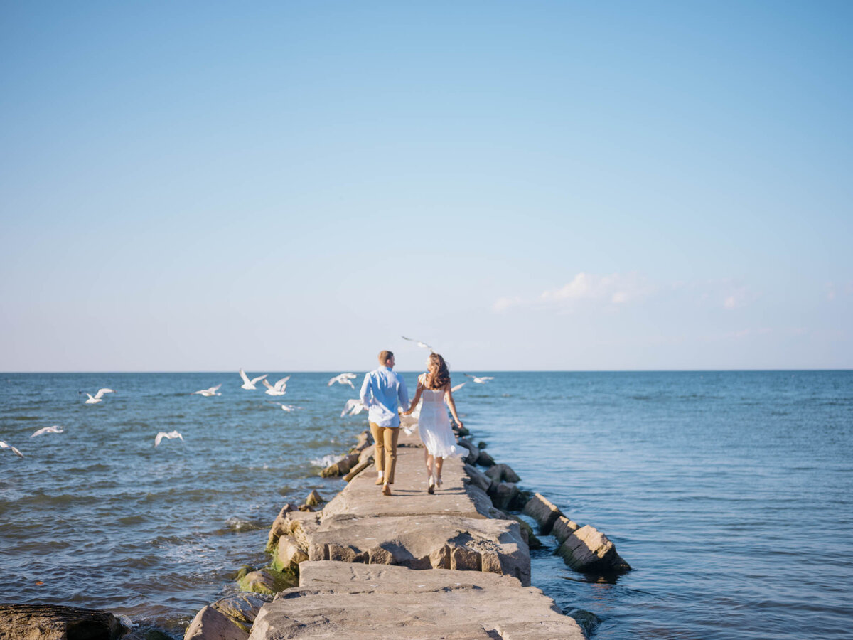 Engagement photo of a couple running hand in hand along a rocky pier in Cleveland, Ohio.