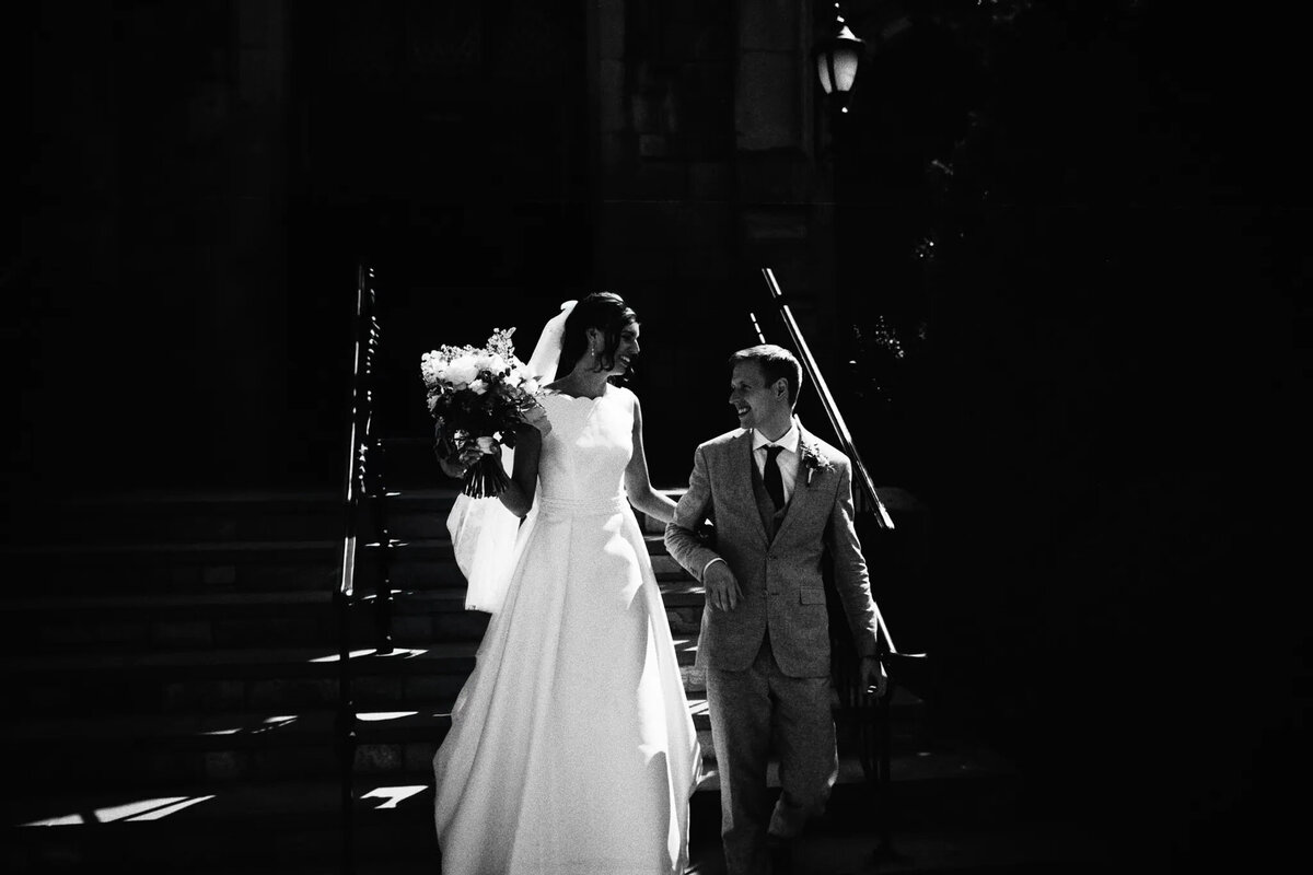 A bride in a long white gown and a groom in a suit walk down outdoor steps together, smiling at each other, with the bride holding a bouquet. Sunlight highlights them against a dark background—captured beautifully by an experienced NJ wedding photographer.