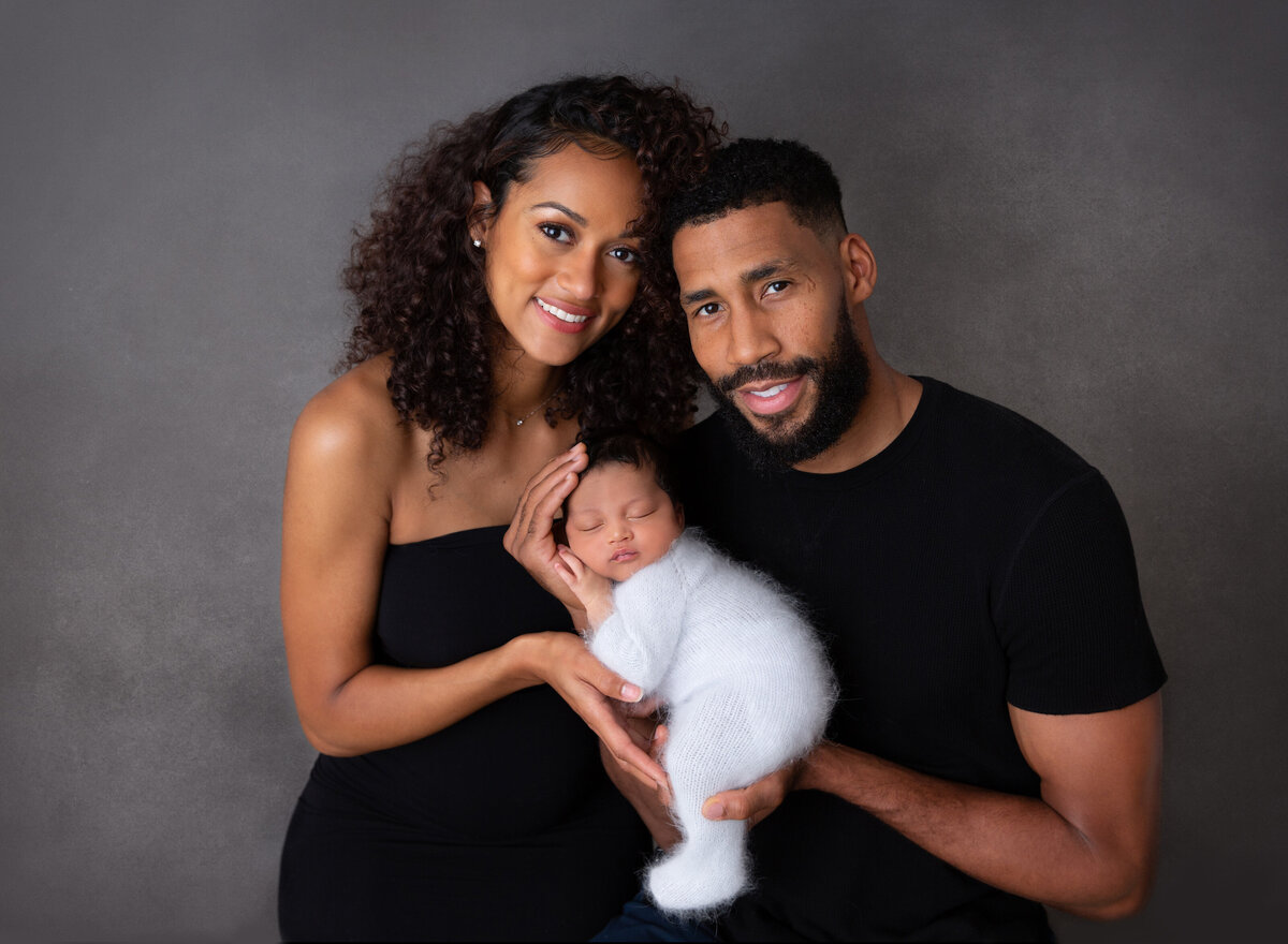 Top newborn photographer Rochel Konik captures family in Brooklyn, NY photography studio.  Mom and Dad holding newborn baby smiling at the camera. Baby is in a flu