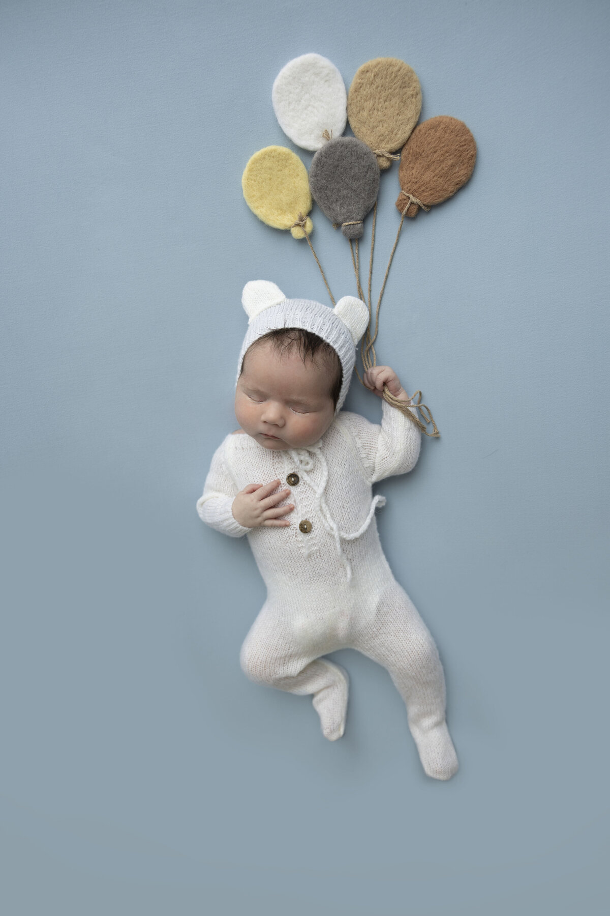 Baby in bear outfit holds onto a bundle of balloons.