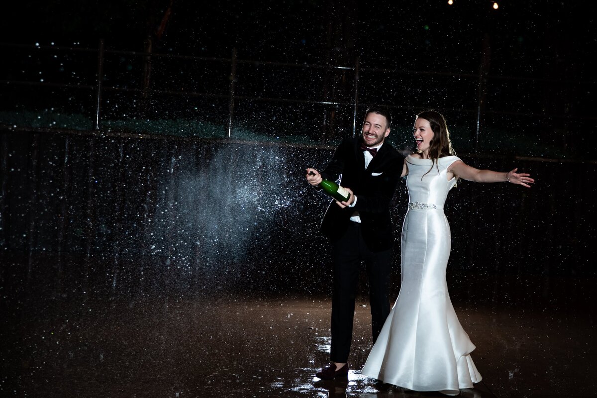 Bride and groom pop a bottle of champagne in the rain after their reception.