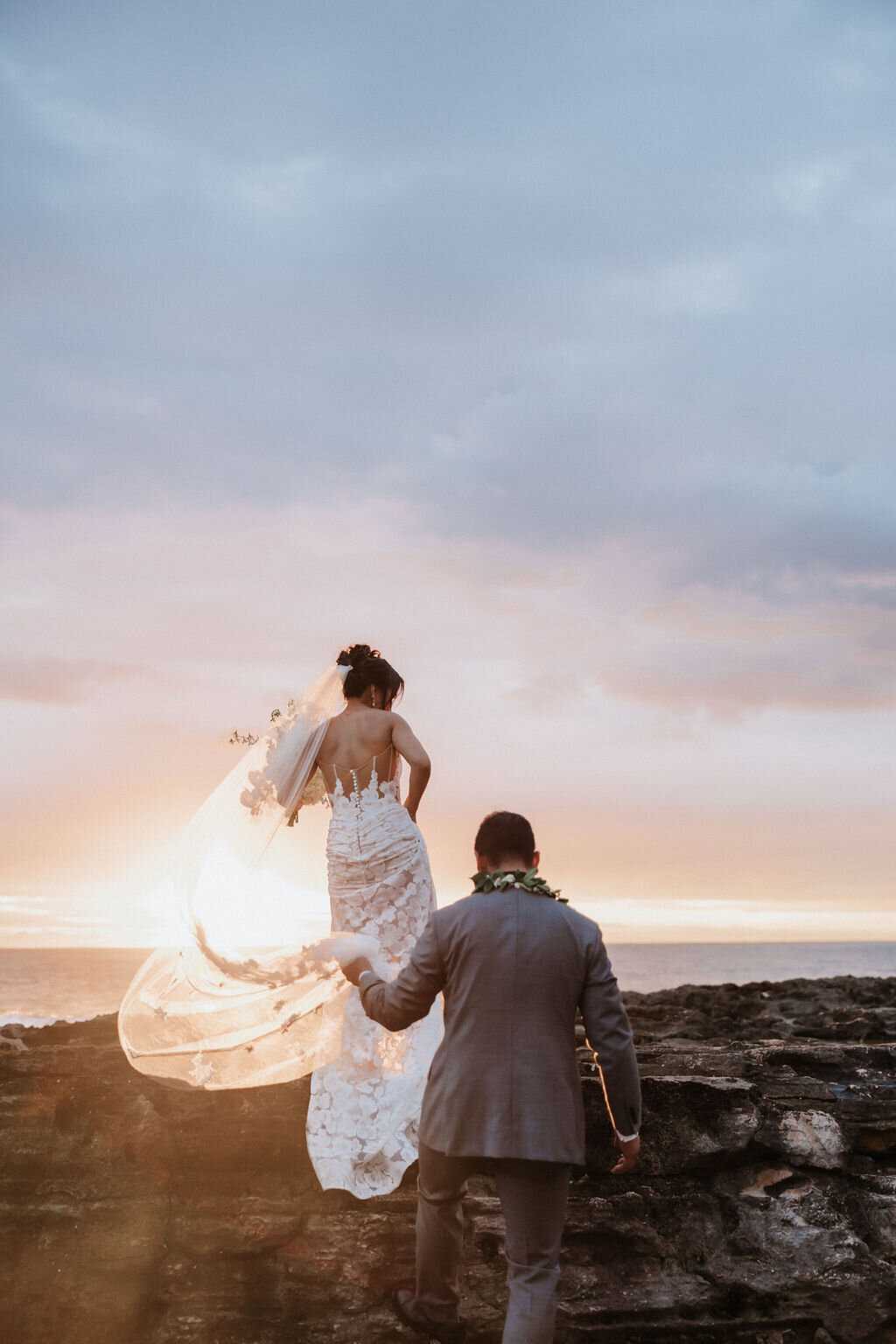 bride and groom walking towards the sunset while groom holds the brides train and veil