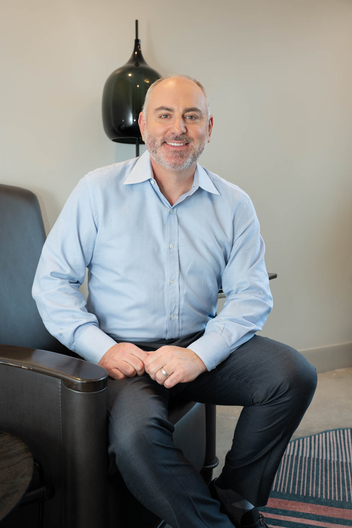 An image of a men wearing a blue shirt and dark pants, sitting on a chair with his hands on one of his legs