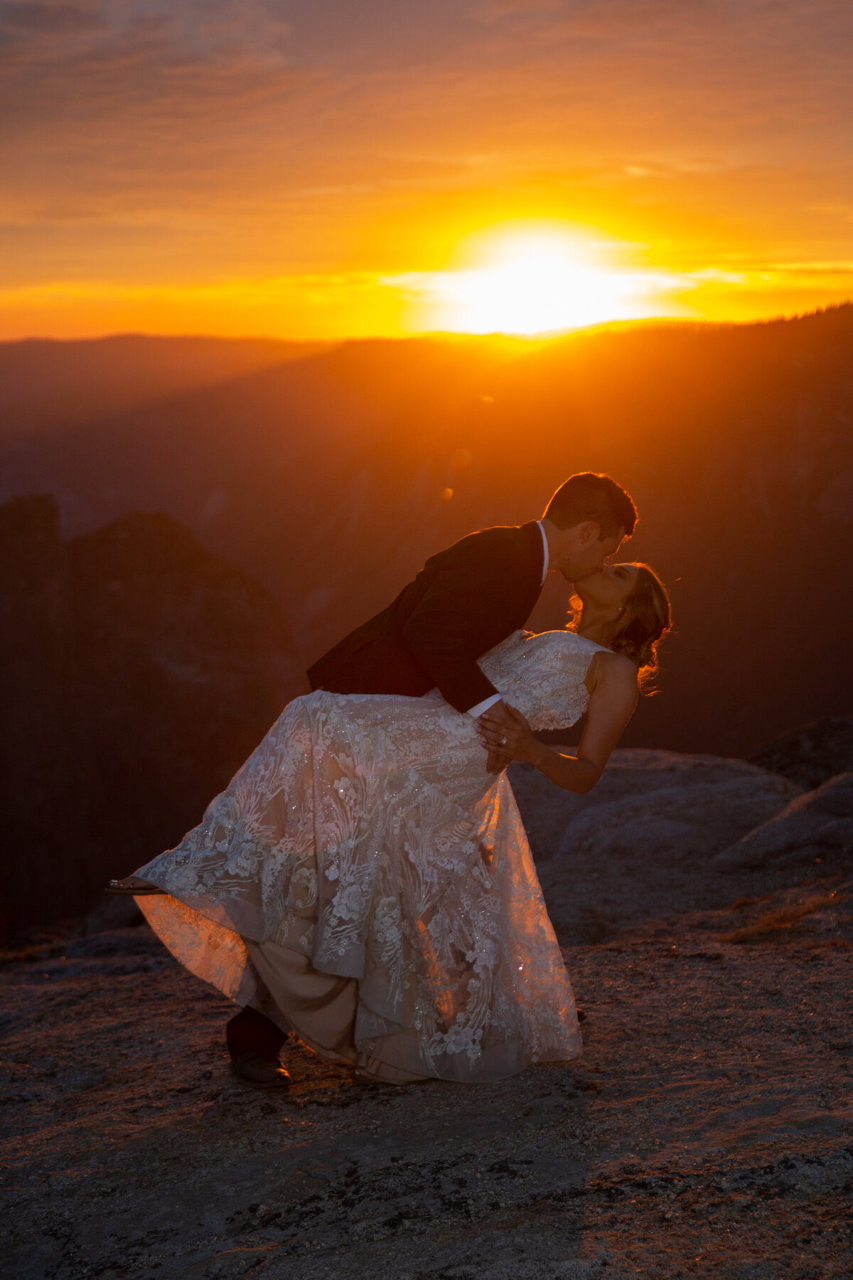 A groom dips his wife and kisses her as the sun turns the sky orange behind them.