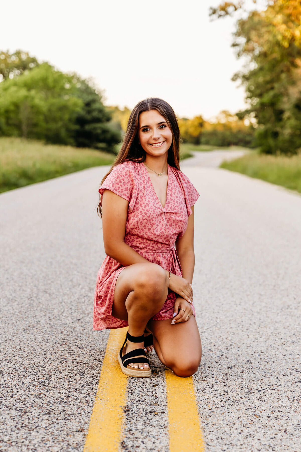 brown haired high school senior girl kneeling on the yellow lines on a back road and smiling for her senior photos