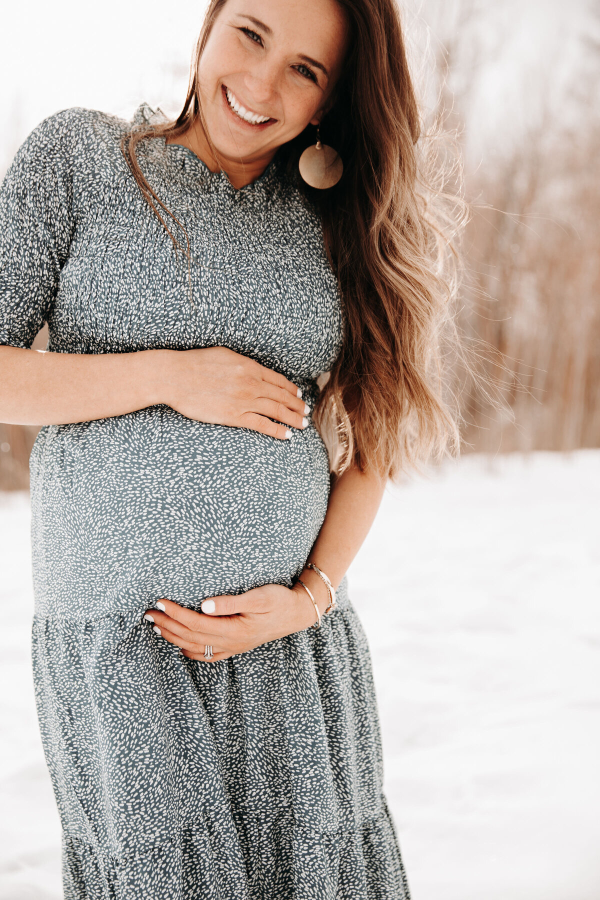 Idaho Falls maternity photographer captures pregnant woman in a blue and white dress holding her baby bump as she smiles in a snowy flied