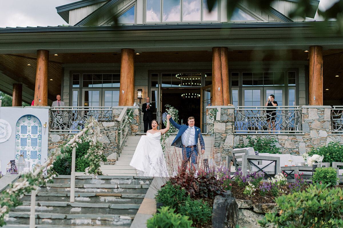 Bride and groom holding hands and cheering while walking down the clubhouse steps during a mountain wedding reception in Cashiers, North Carolina.