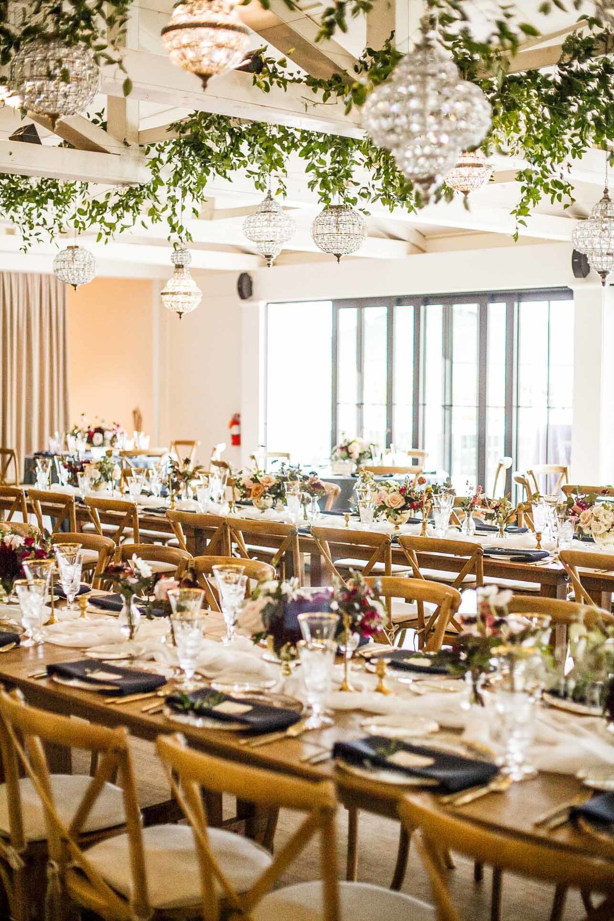 long wooden banquet tables, Vineyard chairs, navy blue napkin, crystal chandeliers, and greenery in ceiling