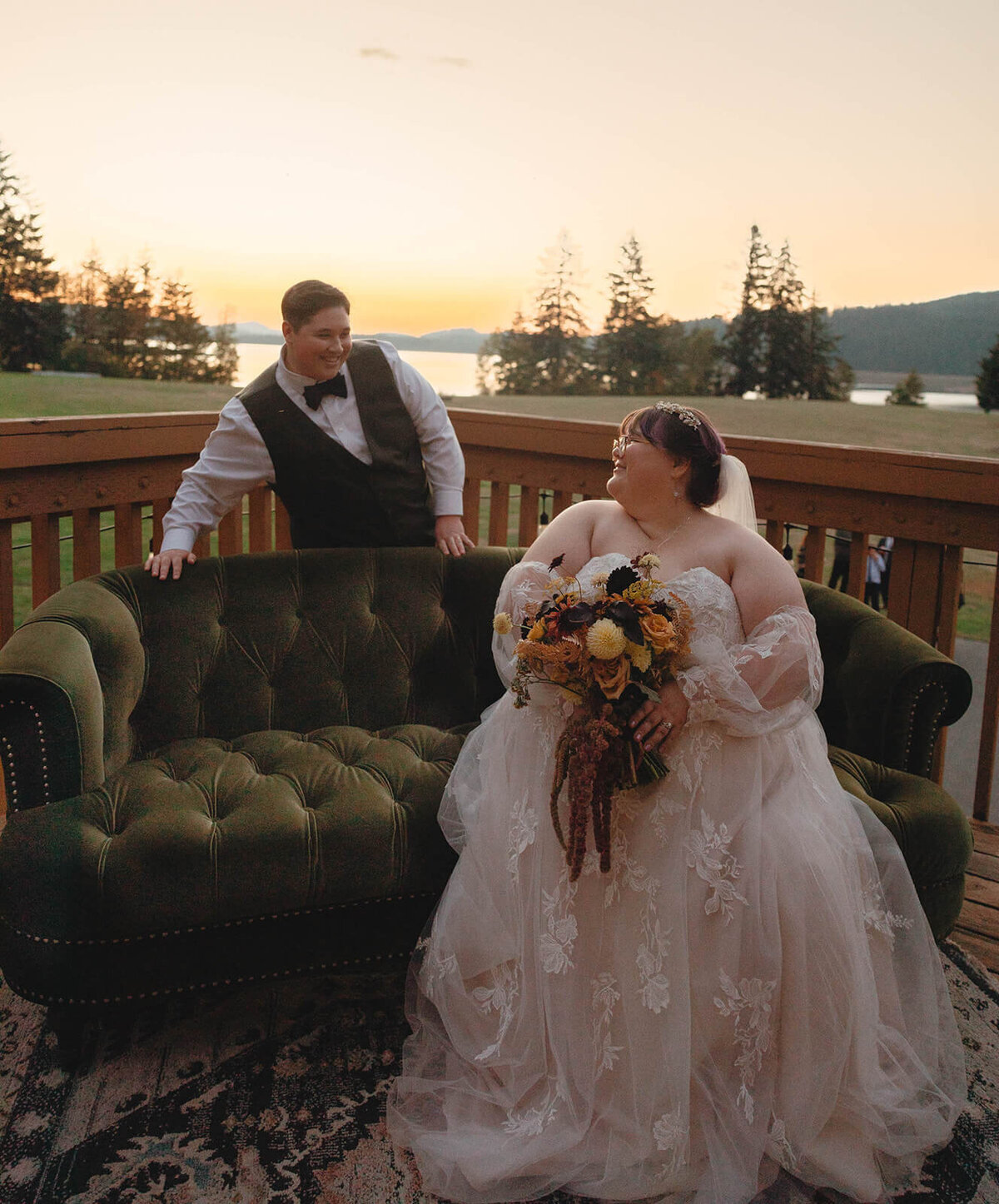 A bride sits on a couch while her wife stands behind the couch and smiles at her at their outdoor wedding.