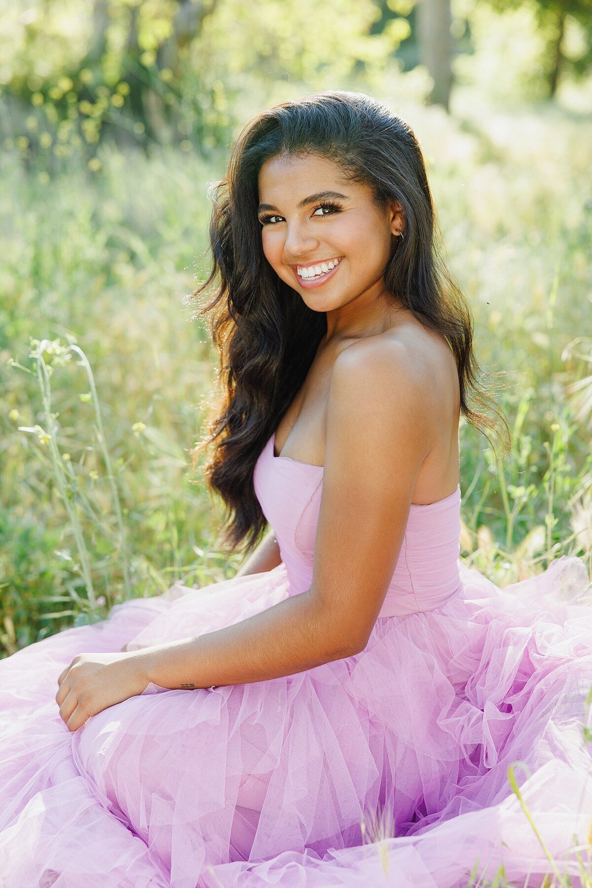 High school senior in a lavender tulle dress sitting in a wildflower field during golden hour, smiling confidently.