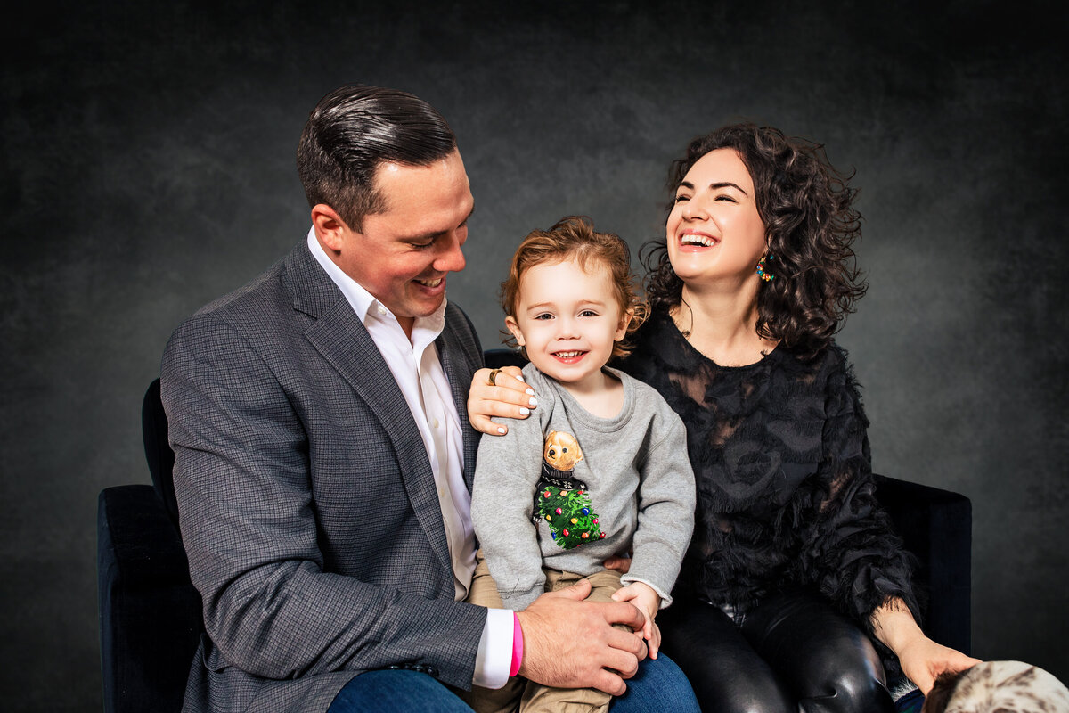 A family of 3 with a small boy in the middle embrace and laugh in the Larger than life photography studio on a vintage dappled grey backdrop.