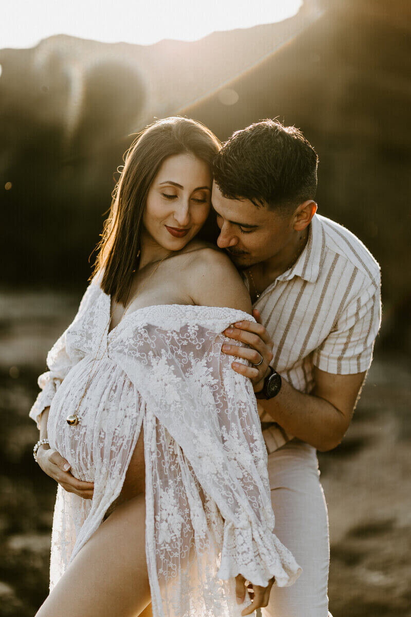 Homme faisant un bisou sur l'épaule de sa femme dénudée. Elle porte uen robe de dentelle blanche laissant apparaitre son ventre bien rond. Photo prise à la plage en Vendée, par Laura, photographe grossesse.