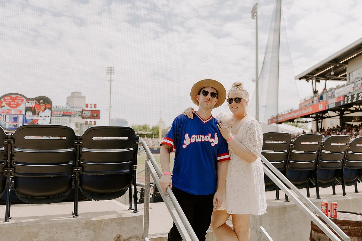 The bride, wearing a lace mini dress embraces the groom wearing a blue Nashville Sounds jersey with a fedora. The bride and groom stand among the seats at the Nashville Sounds Stadium for their baseball wedding at First Horizon Park.
