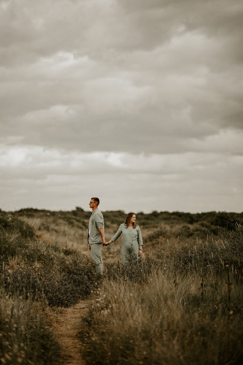 Couple se tenant la main, habillé en vert et regardant dans des directions opposés. Shooting photo grossesse dans les dunes en vendée.