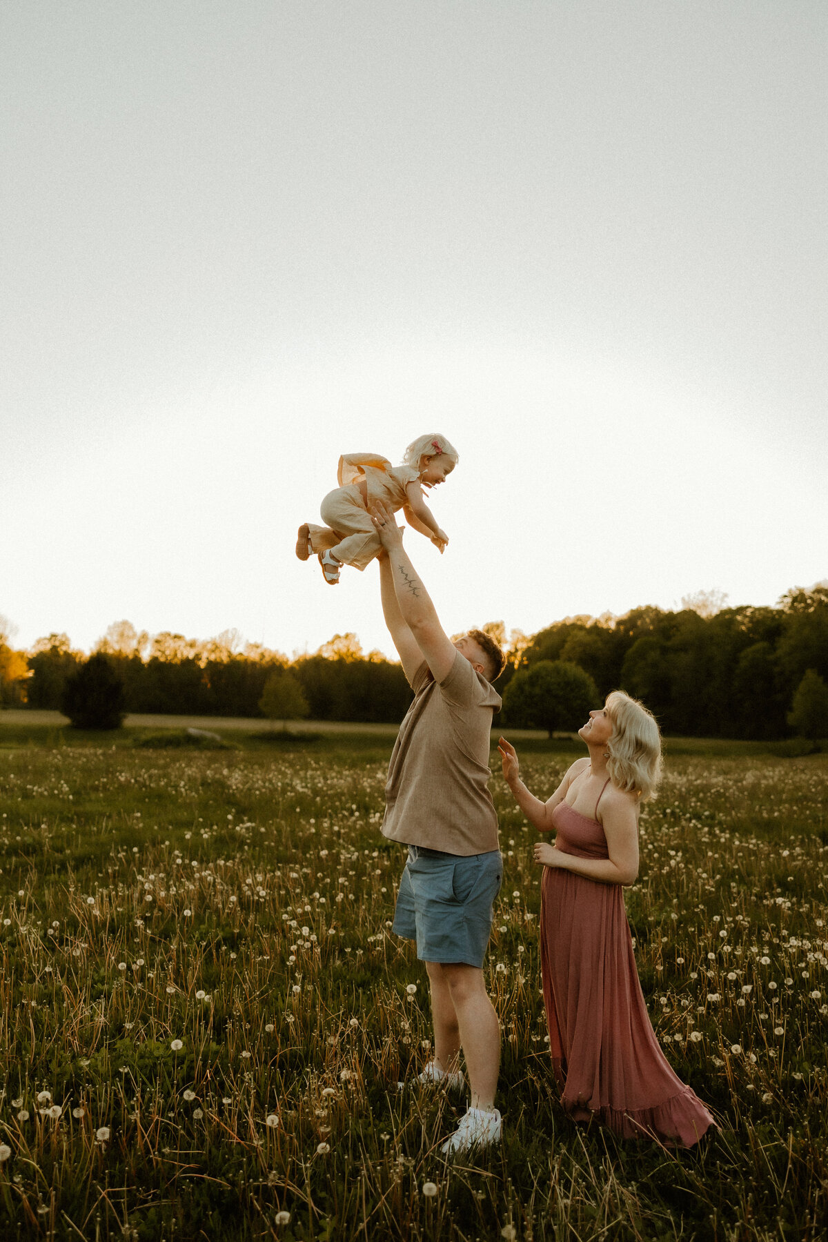 candid photo at sunset of family, dad throwing child in air while mom watches at golden hour