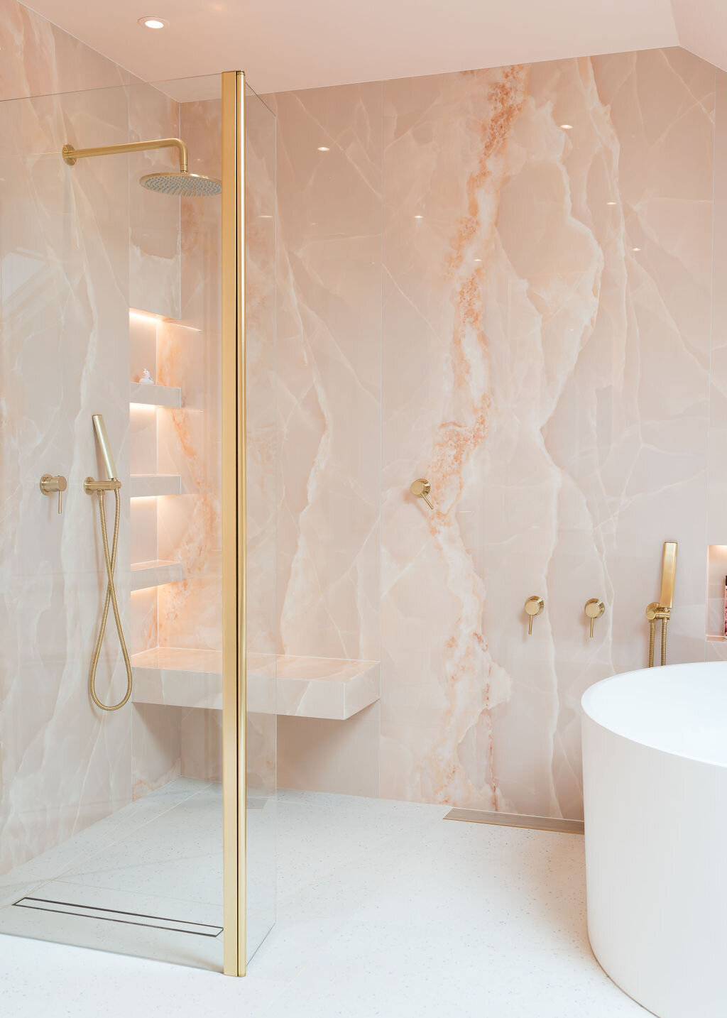 Luxurious bathroom featuring a freestanding round bathtub, gold fixtures, and pink marble walls. A glass-enclosed walk-in shower with built-in shelving is to the left. The room has a clean, modern aesthetic.