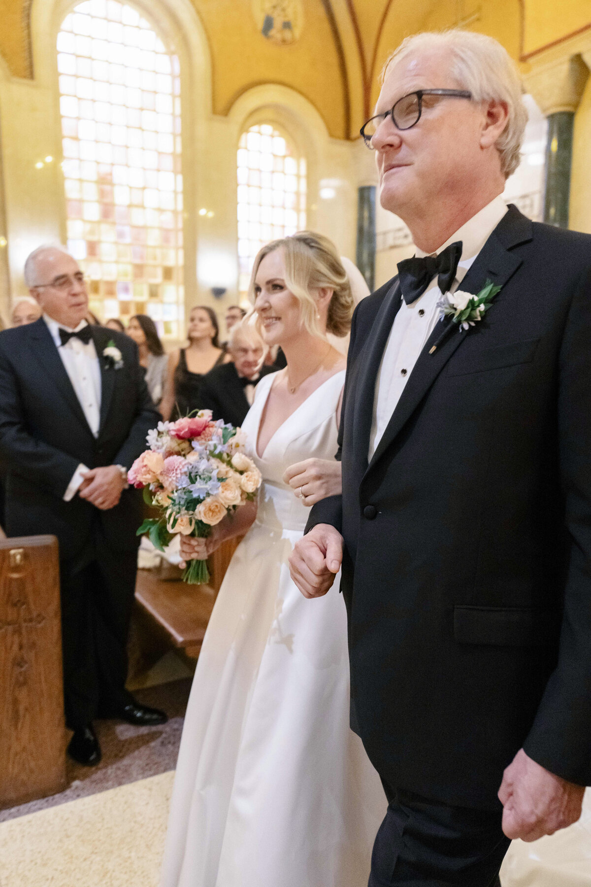 A bride in a white dress holds a bouquet of flowers and walks down the aisle with an older man in a tuxedo. Guests in formal attire watch from the pews in a decorated venue with large stained glass windows.