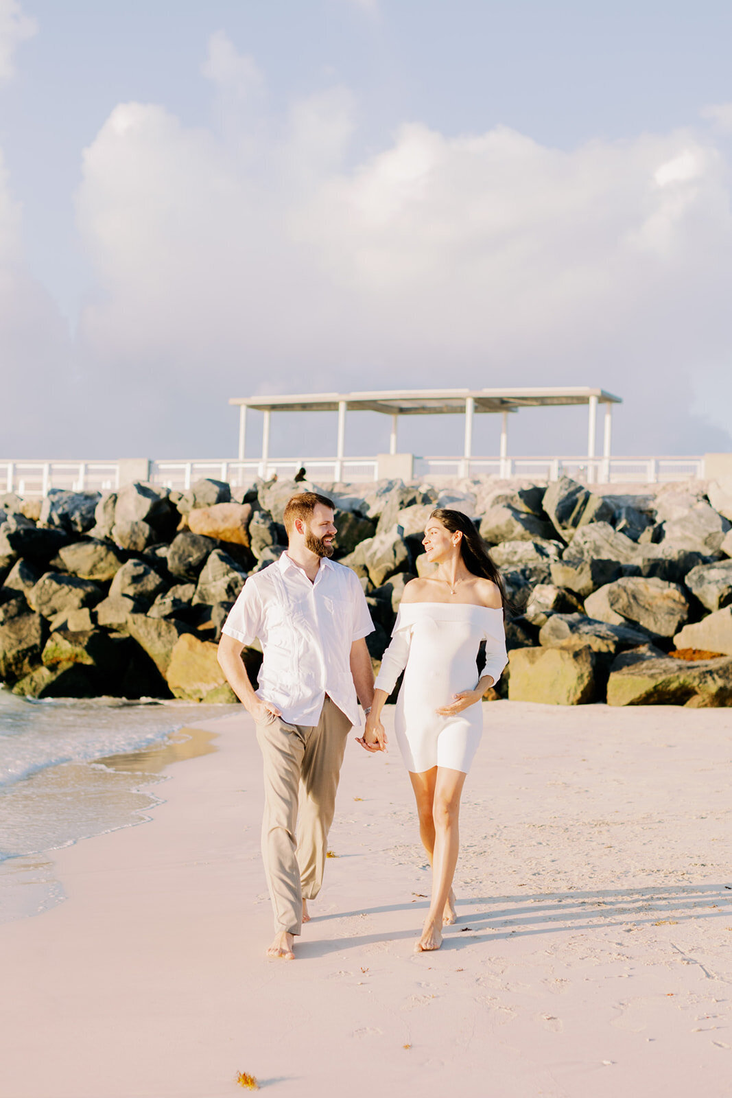 A couple walks barefoot along the sandy shoreline, holding hands and sharing a joyful moment during their maternity session in Miami, Florida. Captured by Claudia Amalia, Miami maternity photographer.