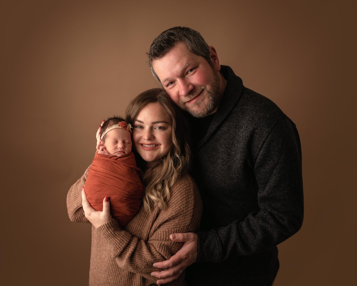 Newborn with mom and dad for studio portrait. Mom and Dad smiling at the camera and baby sleeping.