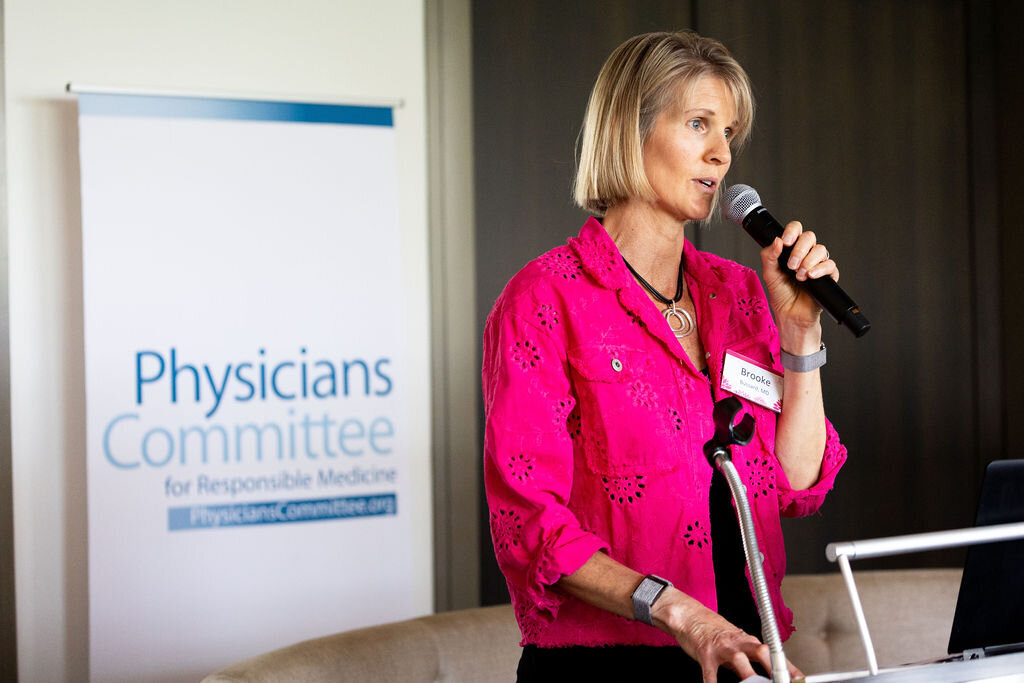 A woman with a pink jacket speaks into a microphone with a "Physicians Committee" sign behind her.