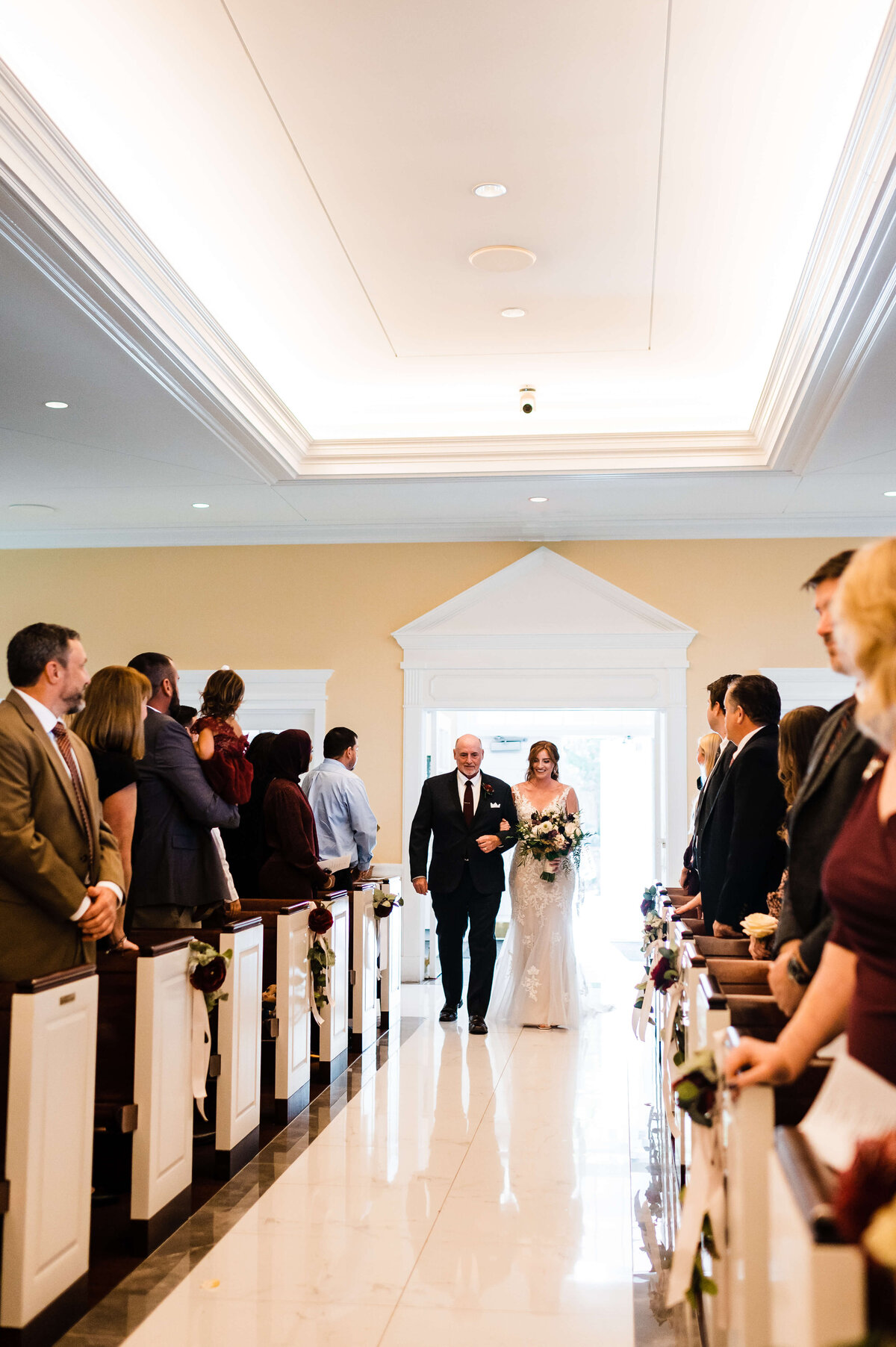 Father of the bride holds his daughter as they walk down the aisle together at a Chapelle wedding in Shenandoah national park with the guests in pews standing in respect of the bride