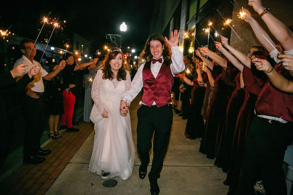 A bride and groom stand smiling, surrounded by their wedding party holding sparklers in celebration. Everyone is dressed formally and appears joyful.