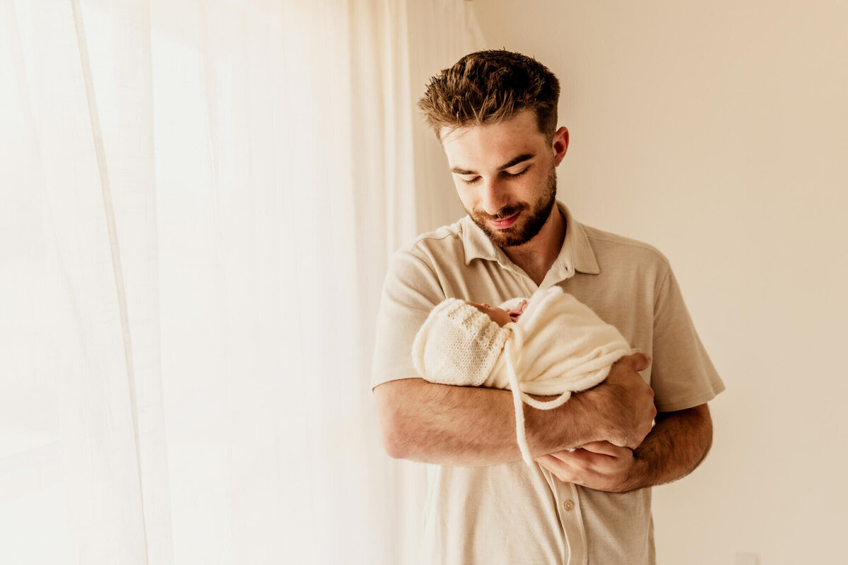 A father wearing a white collared shirt smiles down at his baby girl who is swaddled in his arms in Yukon, Oklahoma.