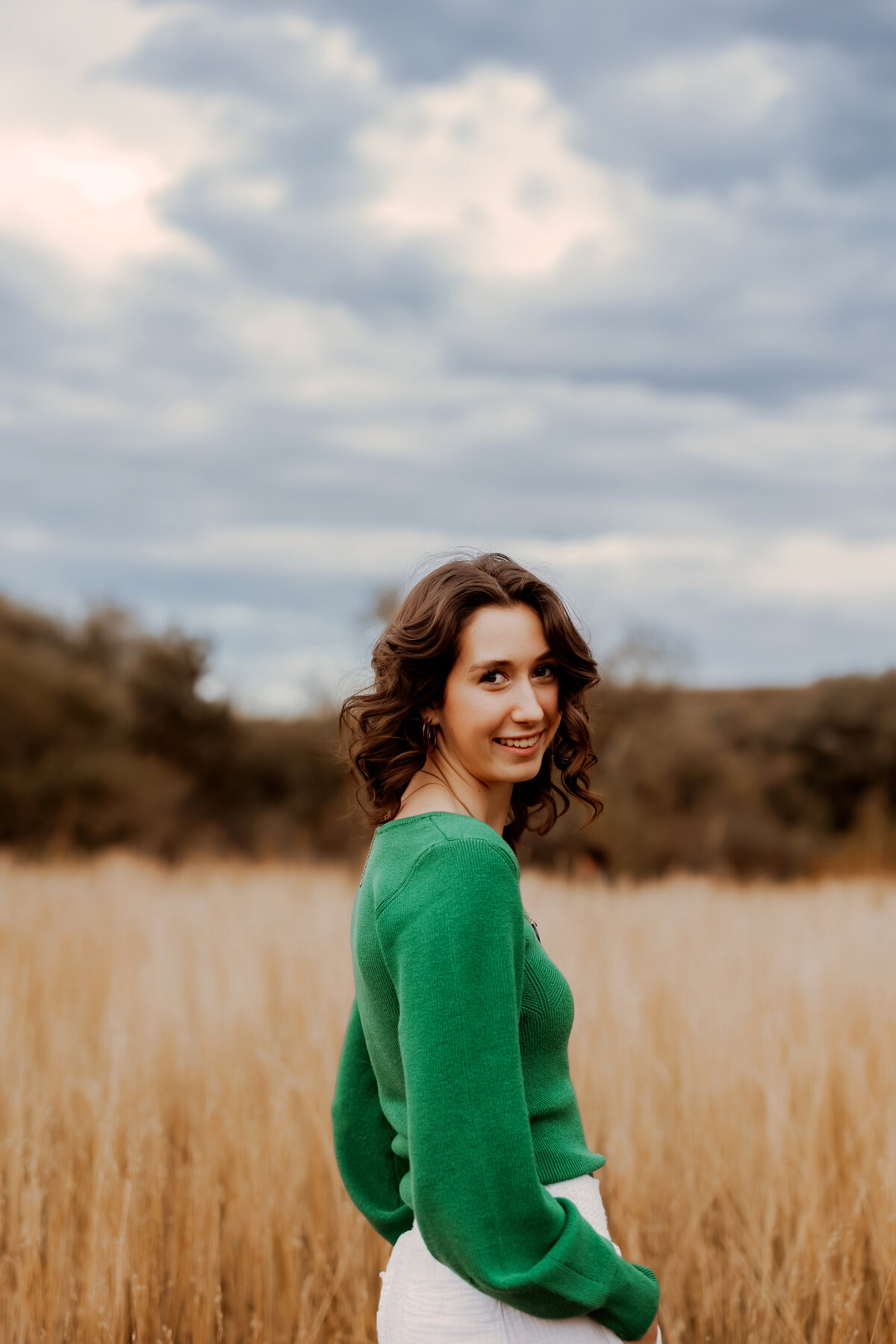High School senior girl in a long sleeve green shirt standing in a golden field with blue skies