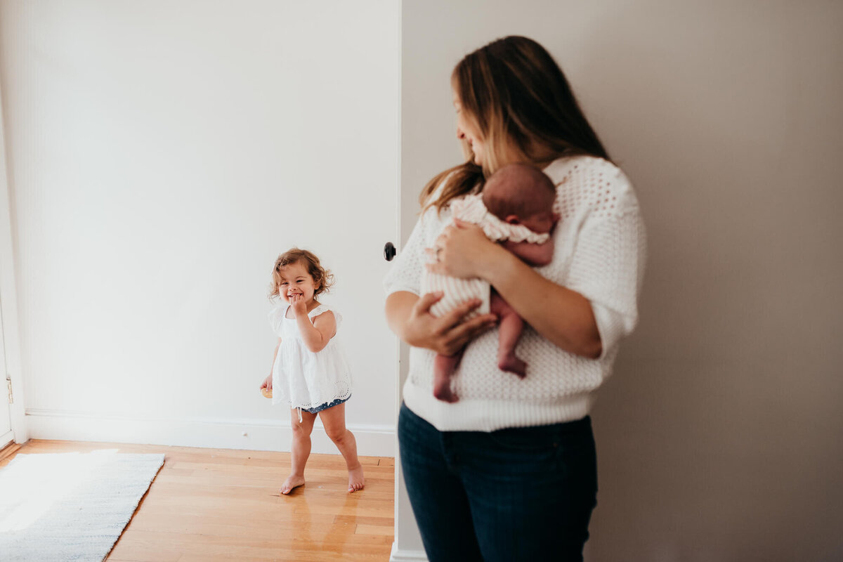 A mother holds her newborn daughter as she looks on at her older daughter who is eating a cracker around the corner