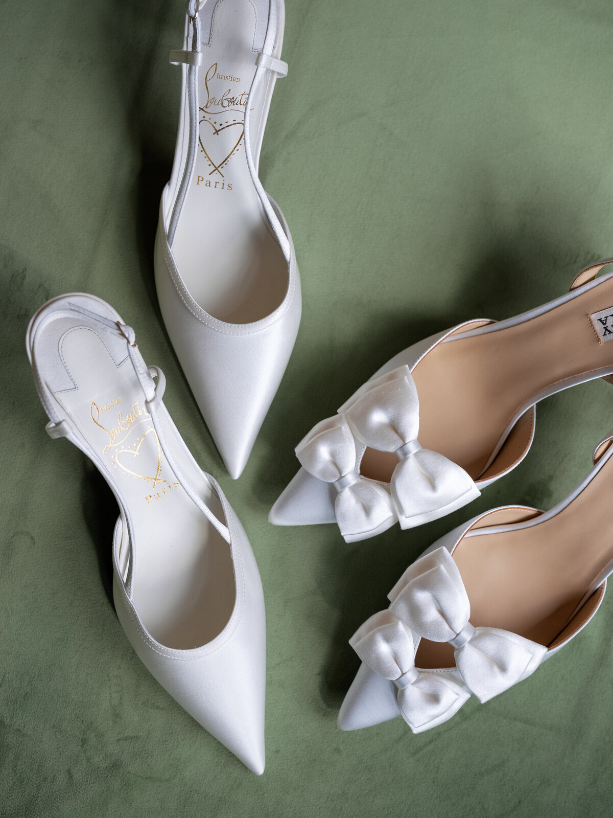 Flat lay of white Louboutin bridal shoes and satin bow heels displayed on a green backdrop for a luxury fall wedding at Castle Ladyhawke in Tuckasegee, NC.