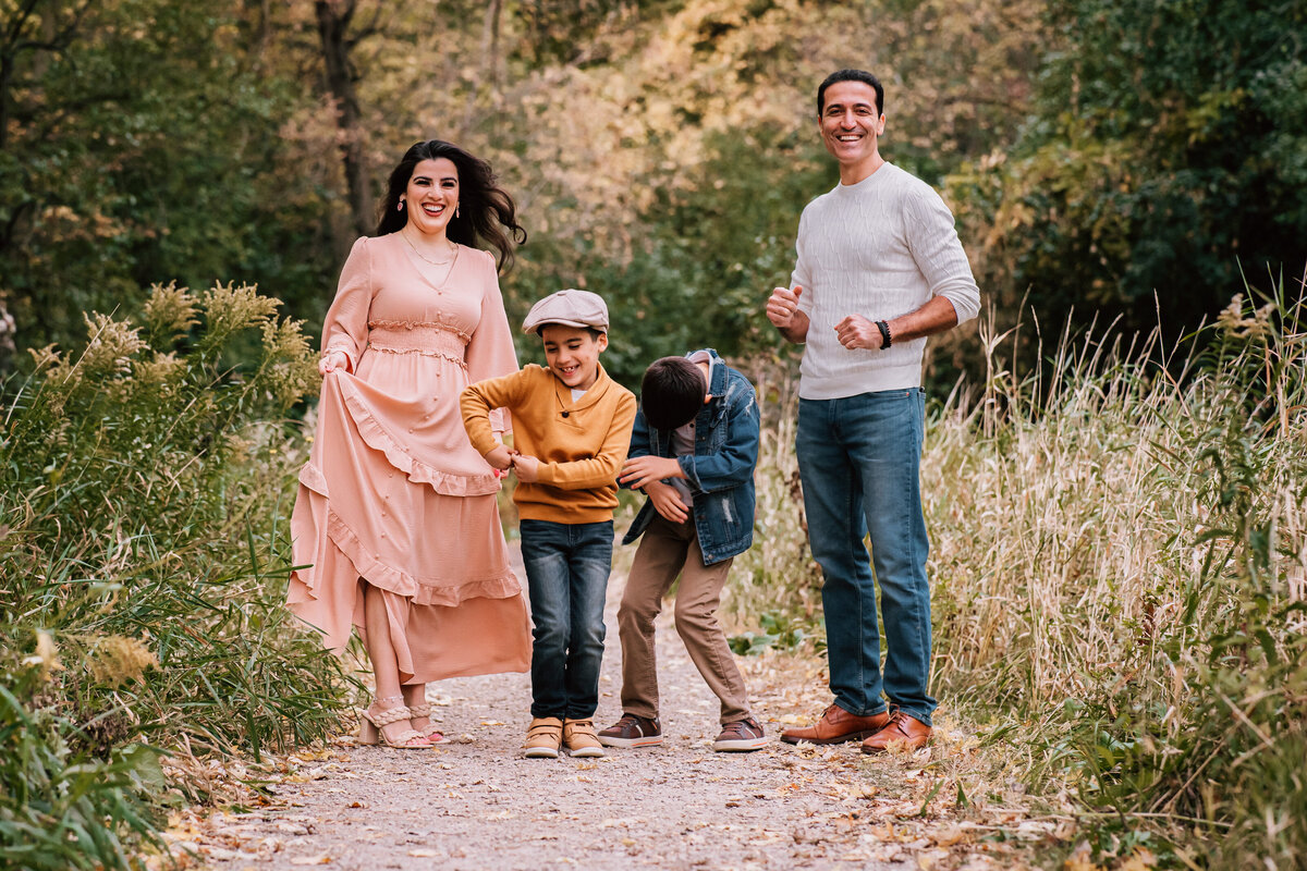 A family of four walking along a tree-lined path. The mother in a flowing pink dress, father in a white sweater, and two boys in casual clothes are all smiling and interacting joyfully as they walk together.