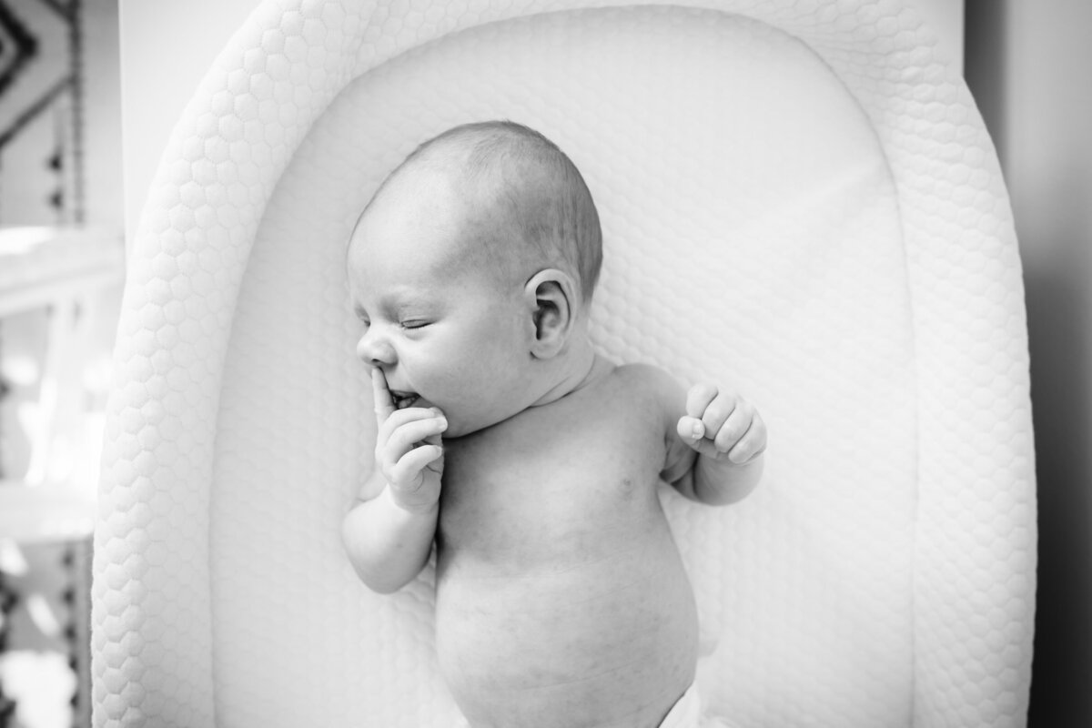 A black-and-white photograph of a newborn baby lying on a white cushioned surface, with their eyes closed and one hand near their mouth, appearing relaxed and peaceful.