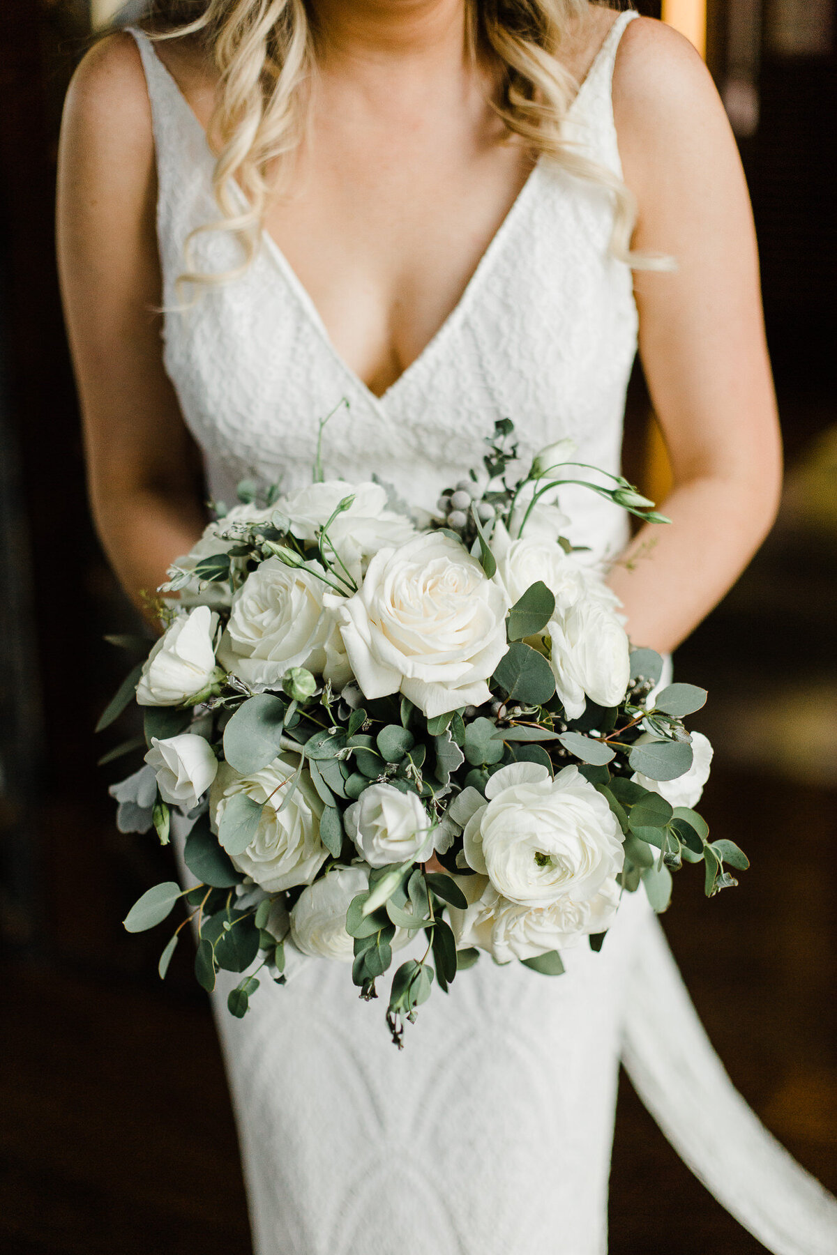Bride carrying all white and greenery loose bouquet with white ranunculus, white lisianthus, and white roses
