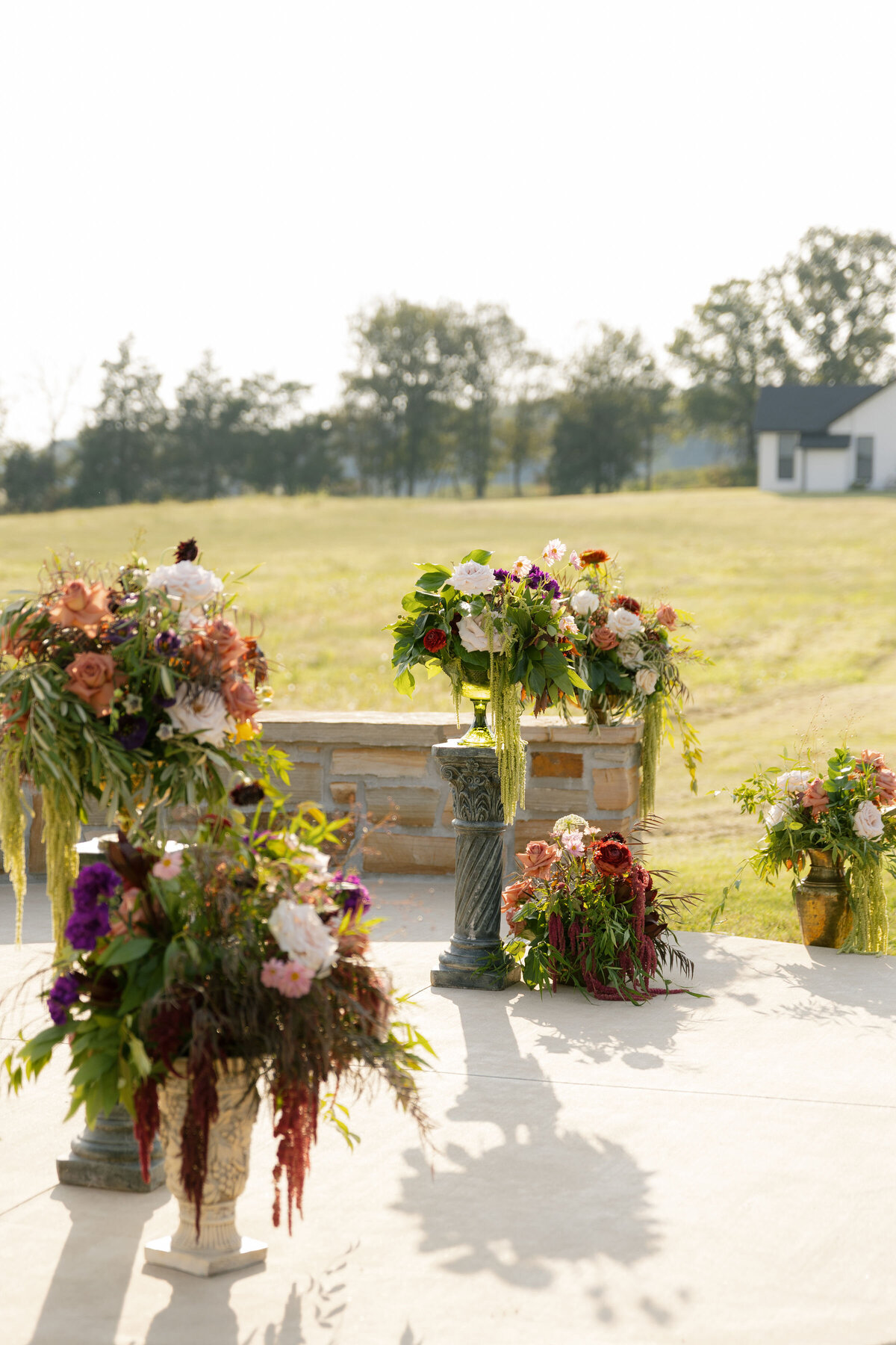 Outdoor ceremony floral installations featuring multiple overflowing arrangements of roses, dahlias, amaranthus, and greenery positioned on stone and pedestal stands in a sunlit field.