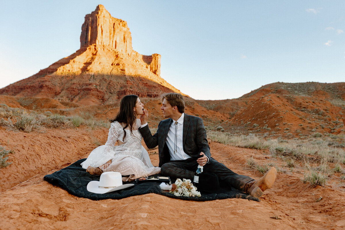 A bride and groom sit having a picnic during their elopement in Moab. the groom is feeding the bride. 